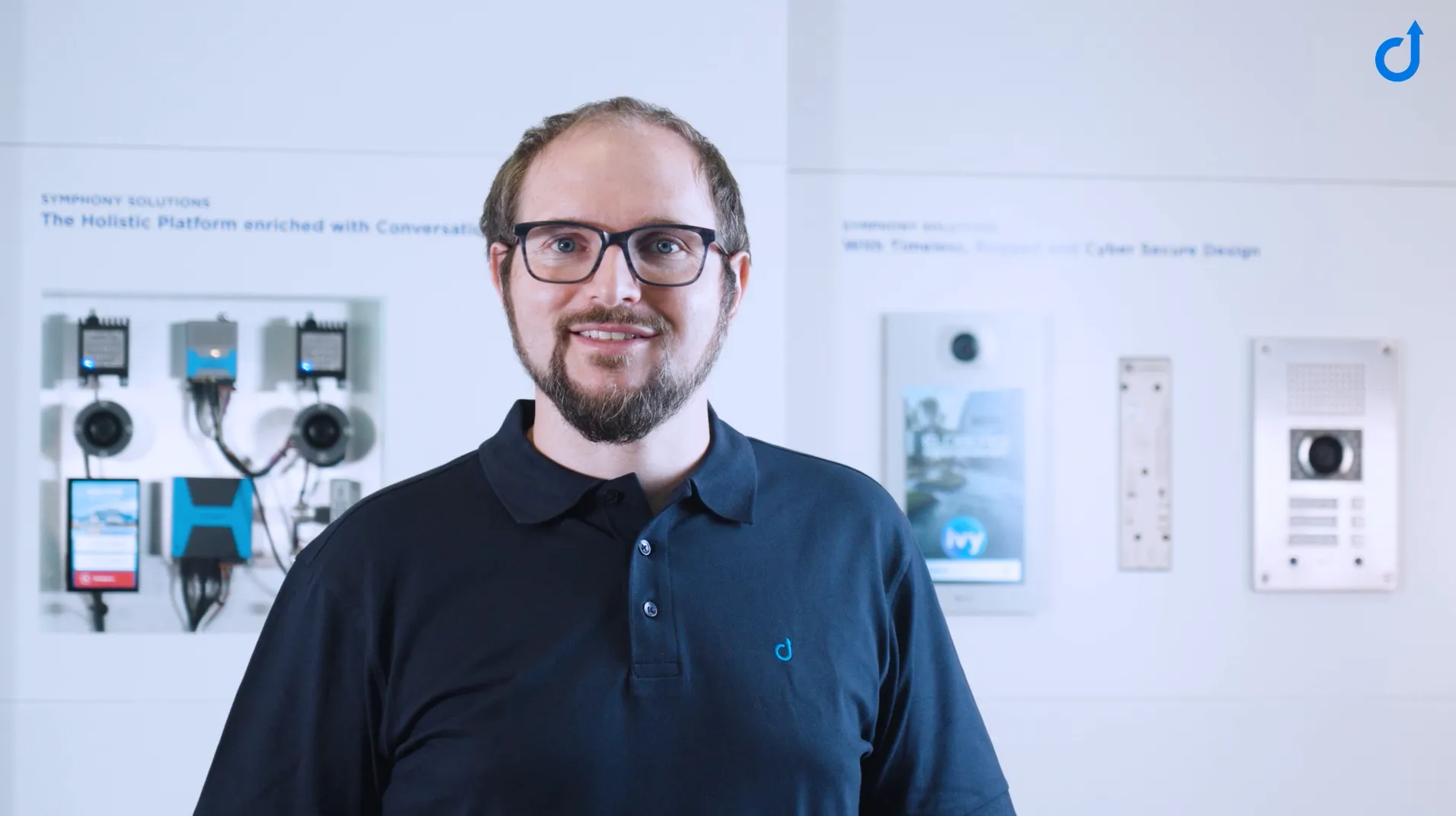 A smiling man in a black polo shirt, standing before a display of diverse intercom and communication systems.